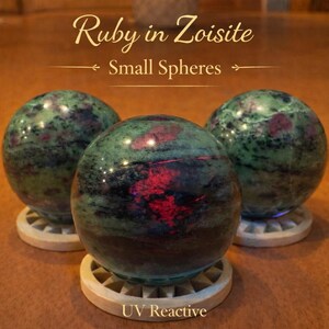 May include: Three small spheres of Ruby in Zoisite, each resting on a light-colored wooden stand. The spheres are primarily green with red and black inclusions. The text "Ruby in Zoisite" and "Small Spheres" is visible. The spheres are UV reactive.