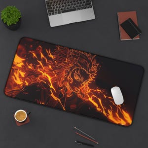 Puede incluir: Una alfombrilla de escritorio negra grande con un diseño de personaje de anime en llamas con llamas naranjas y amarillas. Un ratón de ordenador blanco descansa sobre la alfombrilla, junto a un portátil, una taza de café y artículos de papelería.