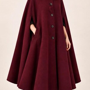 May include: A deep burgundy wool-blend cape coat featuring a structured collar, a front button closure with four visible dark buttons, and side arm slits. The garment has a flared, A-line silhouette that extends to the mid-calf, paired with black trousers and black ankle boots.
