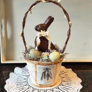 Puede incluir: Una cesta decorativa de Pascua con un conejito de chocolate, huevos moteados y adornos florales. La cesta tiene un asa tejida adornada con capullos blancos. La base es una maceta blanca con una ilustración de zanahorias, sobre un tapete de encaje blanco.