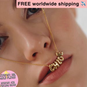 Puede incluir: Un collar chapado en oro con el nombre "CHLOE" escrito en letras de burbujas. El collar se lleva delante de la cara de una persona. El exterior es chapado en oro de 18 quilates y el interior es de plata de ley 925.