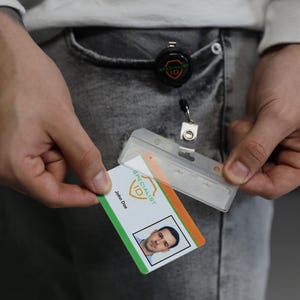 Puede incluir: Un portatarjetas de pl&aacute;stico transparente unido a un carrete retr&aacute;ctil negro con el logotipo de Specialist ID. La tarjeta presenta un fondo blanco con detalles en verde y naranja, el logotipo de Specialist ID, el nombre John Doe y una fotograf&iacute;a de retrato.