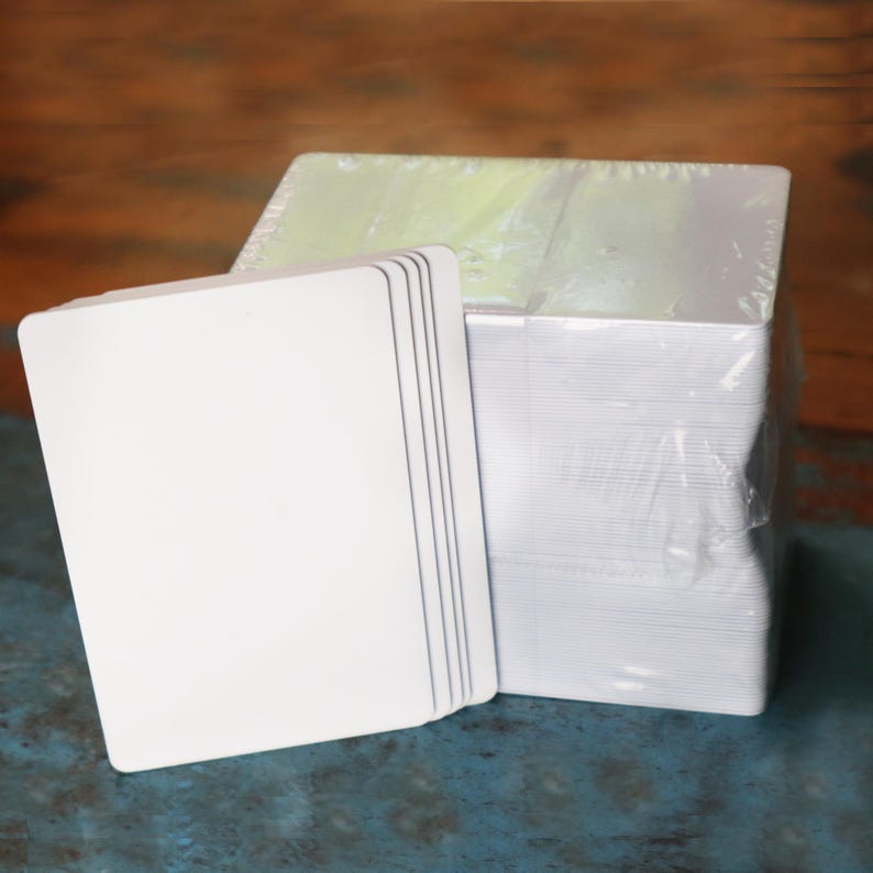 May include: A stack of white, square cards with rounded corners, next to a larger stack of similar cards wrapped in clear plastic. The cards have a smooth, clean surface, and are stacked neatly. The background is a mix of blue and brown.