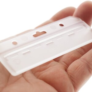 Puede incluir: Un portatarjetas rectangular de pl&aacute;stico esmerilado semitransparente, con una ranura central para clip y dos orificios circulares a los lados. El soporte tiene un dise&ntilde;o r&iacute;gido y moldeado con un acabado mate claro y una superficie lisa.