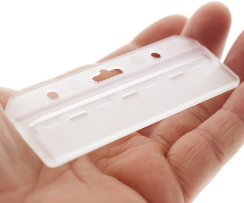 Puede incluir: Un portatarjetas rectangular de pl&aacute;stico esmerilado semitransparente, con una ranura central para clip y dos orificios circulares a los lados. El soporte tiene un dise&ntilde;o r&iacute;gido y moldeado con un acabado mate claro y una superficie lisa.