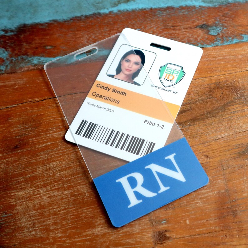 Puede incluir: Una tarjeta de identificaci&oacute;n de pl&aacute;stico blanco con una foto de una mujer, el texto "Cindy Smith Operations Since March 2021 Print 1-2" y un c&oacute;digo de barras. La tarjeta est&aacute; en un porta-tarjetas de pl&aacute;stico transparente con una etiqueta azul que dice "RN".