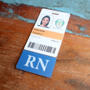 Puede incluir: Una tarjeta de identificaci&oacute;n de pl&aacute;stico blanco con una foto de una mujer, el texto "Cindy Smith Operations Since March 2021", un c&oacute;digo de barras y el texto "Print 1-2". La tarjeta tiene un fondo azul con el texto "RN" en blanco.