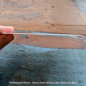 May include: A close-up of a white, rigid sheet of material held vertically. The sheet is held by a hand and shows no signs of bending or flexing. The caption below the sheet reads "Anything But Flimsy - Shown Held Vertically with Nearly No Bend".