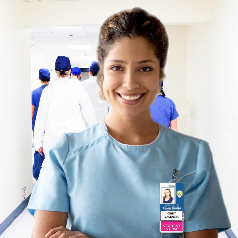May include: A smiling woman wearing a light blue scrub top and a name tag that reads "General Hospital, Cindy Valencia, Student Nurse". She is standing in a hospital hallway with other medical professionals in the background.
