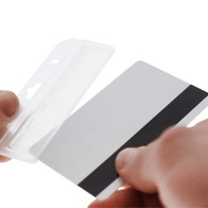 Puede incluir: Un portatarjetas rectangular de pl&aacute;stico transparente con una ranura superior para cord&oacute;n, mostrado mientras se inserta una tarjeta de pl&aacute;stico blanca est&aacute;ndar que presenta una banda magn&eacute;tica negra central para identificaci&oacute;n o acceso.
