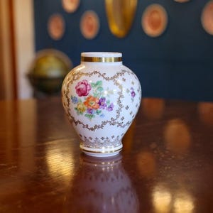 以下が含まれることがあります： 花柄の白いセラミック花瓶。 金色の縁と底があり、カラフルな花と金色のアクセントの模様があります。 花瓶の高さは約15cmです。