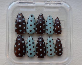 Pode incluir: Um conjunto de dez unhas postiças em forma de amêndoa. As unhas alternam entre uma cor castanha escura e azul claro, cada uma adornada com pequenas bolinhas castanhas escuras espaçadas uniformemente. As unhas são exibidas numa caixa de plástico transparente.