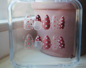 Pode incluir: Um conjunto de unhas postiças em uma caixa de plástico transparente. As unhas são ovais e apresentam um desenho de bolinhas vermelhas sobre uma base transparente ou rosa. A caixa é transparente e o fundo é rosa desfocado.