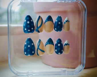 Pode incluir: Um conjunto de unhas postiças numa caixa de plástico transparente. As unhas apresentam vários designs, incluindo azul marinho com bolinhas brancas, bege com detalhes azuis e bege com um desenho de onda azul. As unhas estão numa caixa de plástico transparente.