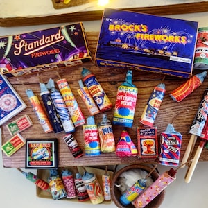May include: A collection of miniature fireworks, including boxes labeled "Brock's Fireworks" and "Standard Fireworks." Various firecrackers and bottle rockets are arranged on a wooden surface, with colorful labels and designs. A small bowl is also present.