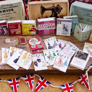 Puede incluir: Una colección de artículos de costura en miniatura, que incluyen libros, cajas y portadas de patrones Vogue. Los artículos están dispuestos sobre una superficie de madera, con banderas de la Union Jack en el fondo. Los libros incluyen títulos como "Singer Fashion Tailoring".