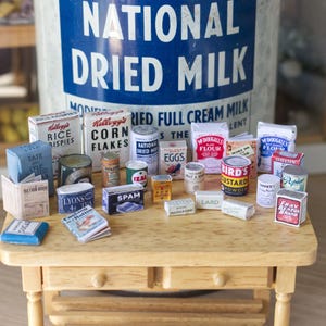 Può includere: Un tavolino in legno in miniatura espone una collezione di generi alimentari in miniatura in stile vintage. Gli articoli includono scatole di cereali, farina e conserve, con etichette come "National Dried Milk" e "Spam".