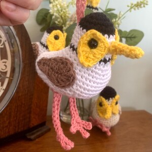 May include: A crocheted bird ornament featuring a white body, brown wings, a black cap, and a yellow face with a pointed beak. The bird has long pink legs and a pink hanging loop. Two additional smaller crocheted bird figures are visible nearby.