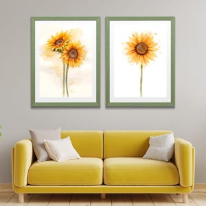 May include: Two framed watercolor prints of sunflowers. One print features three sunflowers, and the other shows a single sunflower. The frames are a muted green, and the background is a neutral color. The artwork is displayed above a yellow sofa.