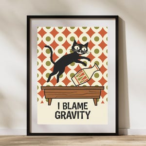 Könnte beinhalten: Gerahmter Kunstdruck mit einer schwarzen Katze, die über einen Holztisch springt und eine Flasche umwirft. Der Hintergrund hat ein Retro-Muster aus orangefarbenen und grünen Kreisen. Der Text "I BLAME GRAVITY" steht darunter.