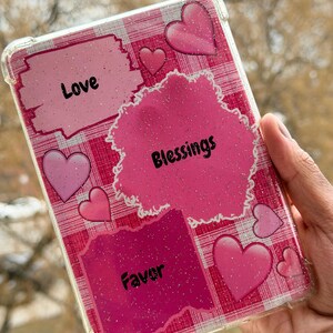 May include: Clear phone case with a pink and white plaid pattern. The case features pink hearts and the words "Love", "Blessings", and "Favor" in black text. The case is held in a hand.