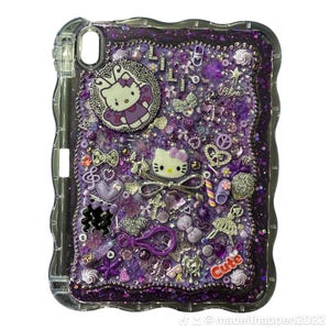 Könnte beinhalten: Eine transparente Tablet-Hülle mit einem Design aus lila Glitzer und Charms. Die Hülle verfügt über verschiedene lila und silberne Verzierungen, darunter Hello Kitty, Schleifen und Herzen. Das Wort "Cute" ist auf der Hülle sichtbar.