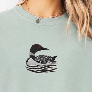 May include: A light green t-shirt with a black and white embroidered loon design. The loon is swimming in water with waves.