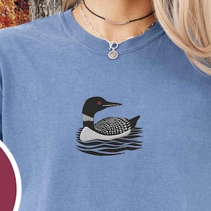 May include: A blue t-shirt with an embroidered loon design. The text "EMBROIDERED COMFORT COLORS 8 COLOR OPTIONS" is on a maroon circle.