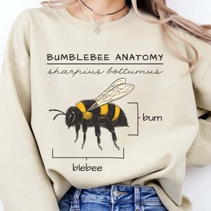 Pu&ograve; includere: Una felpa beige chiaro con una grafica di un bombo e il testo "Bumblebee Anatomy, sharpus bottomus, bum, blebee".