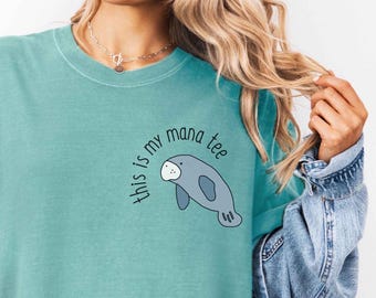Peut inclure: T-shirt turquoise avec l'inscription "this is my mana tee" et un dessin de lamantin gris et blanc. Le t-shirt a un col rond et des manches courtes.