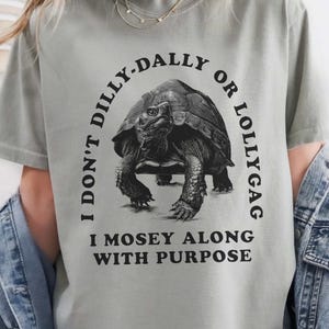 May include: A gray t-shirt with black text that reads 'I don't dilly-dally or lollygag I mosey along with purpose.' A black and white illustration of a tortoise is in the center of the text.