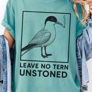 May include: Seafoam green t-shirt with a black square containing a bird illustration smoking a cigarette. The text "LEAVE NO TERN UNSTONED" is below the bird. The word "SEAFOAM" is printed on the left side of the shirt.