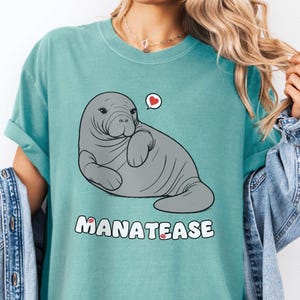 Puede incluir: Una camiseta azul turquesa con un dibujo animado de un manatí con un globo de diálogo en forma de corazón. El texto "MANATEASE" está impreso debajo del manatí.