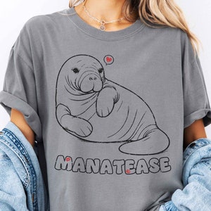 Puede incluir: Camiseta gris con un dibujo de manatí y un corazón encima. El texto "MANATEASE" está impreso debajo en una fuente desgastada. La camiseta tiene un corte relajado.