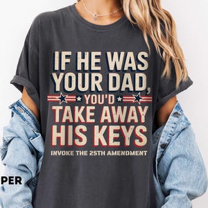Op de afbeelding: Donkergrijs t-shirt met de tekst "IF HE WAS YOUR DAD, YOU'D TAKE AWAY HIS KEYS" in wit, rood en blauw. De zin "INVOKE THE 25TH AMENDMENT" staat eronder. Het woord "PEPPER" staat linksonder.