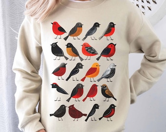 May include: A beige sweatshirt with a colorful pattern of birds. The birds are in various shades of red, orange, yellow, black, and gray. The birds are arranged in rows and columns on the sweatshirt.