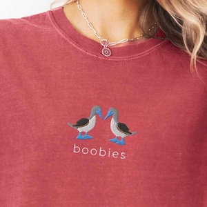 May include: A maroon t-shirt with an embroidered design of two blue-footed boobies facing each other. The word "boobies" is embroidered below the birds. The shirt has a crew neck and short sleeves.