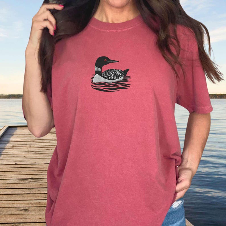 May include: A red t-shirt with a black and white loon graphic on the front. The loon is swimming in water with waves.