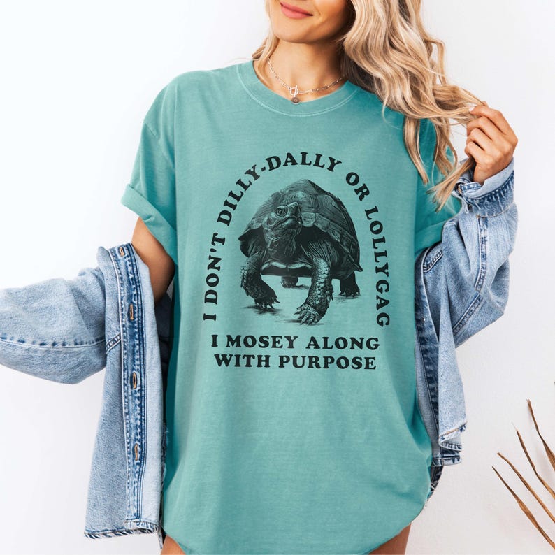May include: A teal blue t-shirt with a graphic of a tortoise and the text "I don't dilly-dally or lollygag I mosey along with purpose".