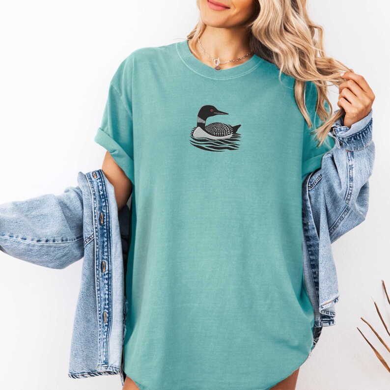 May include: A teal blue t-shirt with a black and white loon graphic on the front. The loon is swimming in water.