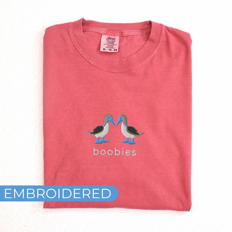 May include: Coral-colored t-shirt with an embroidered design of two blue-footed boobies facing each other. The word "boobies" is embroidered below the birds. The shirt has a Comfort Colors tag and the word "EMBROIDERED" in a blue banner.