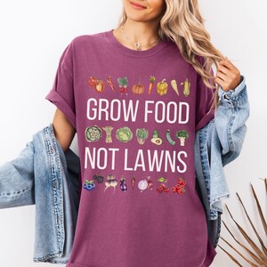 May include: A maroon t-shirt with the text "GROW FOOD NOT LAWNS" in white. The shirt features watercolor illustrations of various vegetables and fruits above and below the text, including tomatoes, carrots, and berries.