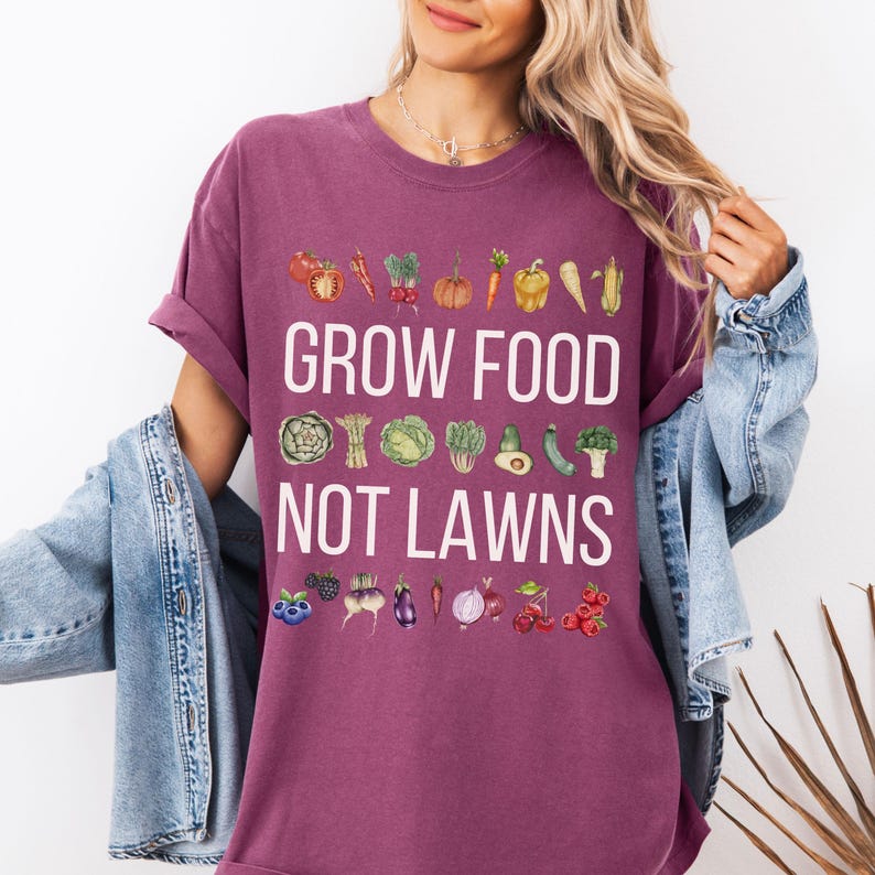 May include: A maroon t-shirt with the text "GROW FOOD NOT LAWNS" in white. The shirt features watercolor illustrations of various vegetables and fruits above and below the text, including tomatoes, carrots, and berries.