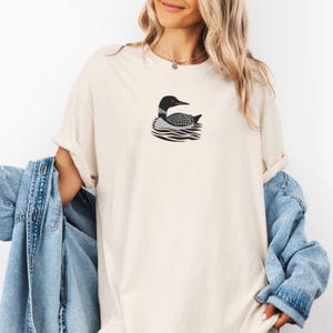 May include: A white t-shirt with a black and white illustration of a loon swimming on the water. The loon has red eyes.