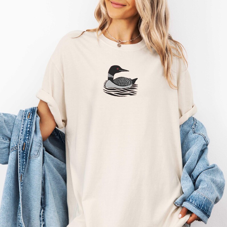 May include: A white t-shirt with a black and white illustration of a loon swimming on the water. The loon has red eyes.