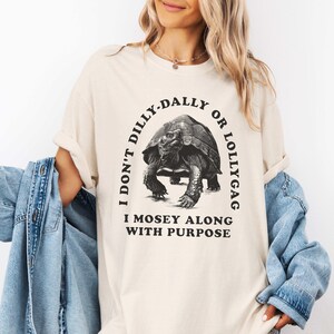 May include: A beige t-shirt with a black graphic of a tortoise and the text "I don't dilly-dally or lollygag I mosey along with purpose".