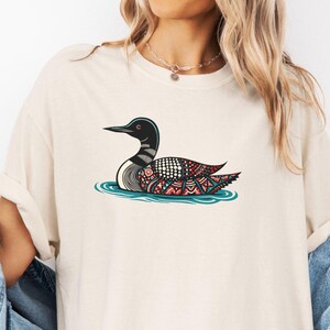 May include: A white t-shirt with a black and white patterned loon design. The loon is swimming in blue water with red and white accents.