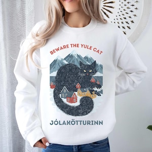 May include: White sweatshirt featuring a graphic of a black cat with yellow eyes, mountains, and colorful houses. Text above reads "BEWARE THE YULE CAT" and below "J&Oacute;LAK&Ouml;TTURINN". The design is a winter holiday theme.