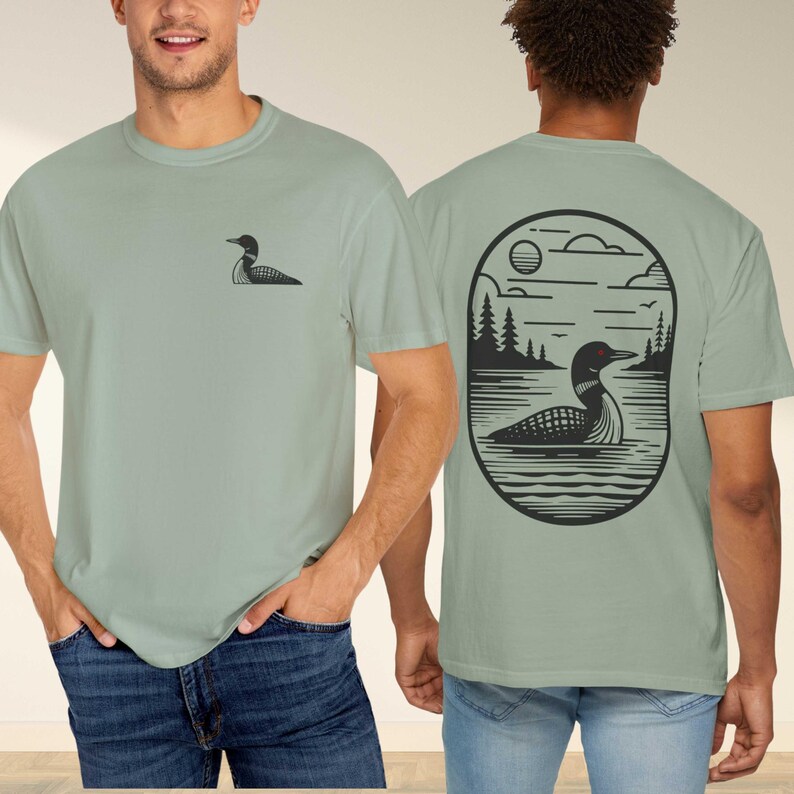 May include: Sage green t-shirts with a black graphic of a loon. One shirt has a small loon graphic on the chest. The other shirt has a larger loon graphic on the back, set within an oval frame with a lake and trees.