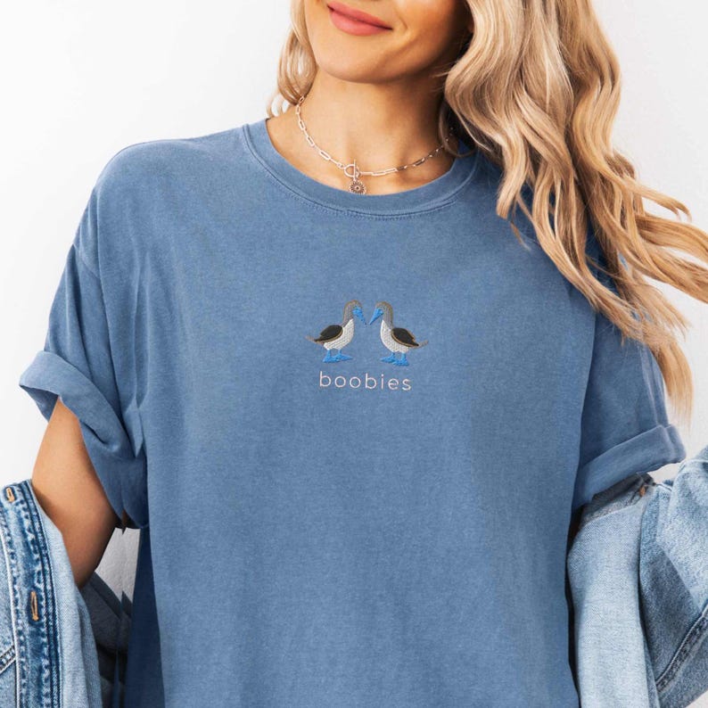 May include: A blue t-shirt with a crew neck and short sleeves. The shirt features an embroidered design of two blue-footed boobies facing each other, with the word "boobies" embroidered below them in a light blue color.